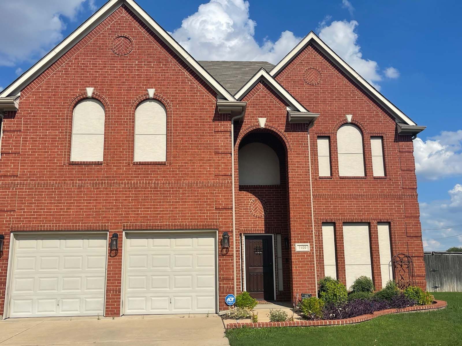 Two-story brick house with covered windows and garage doors, under a blue sky.