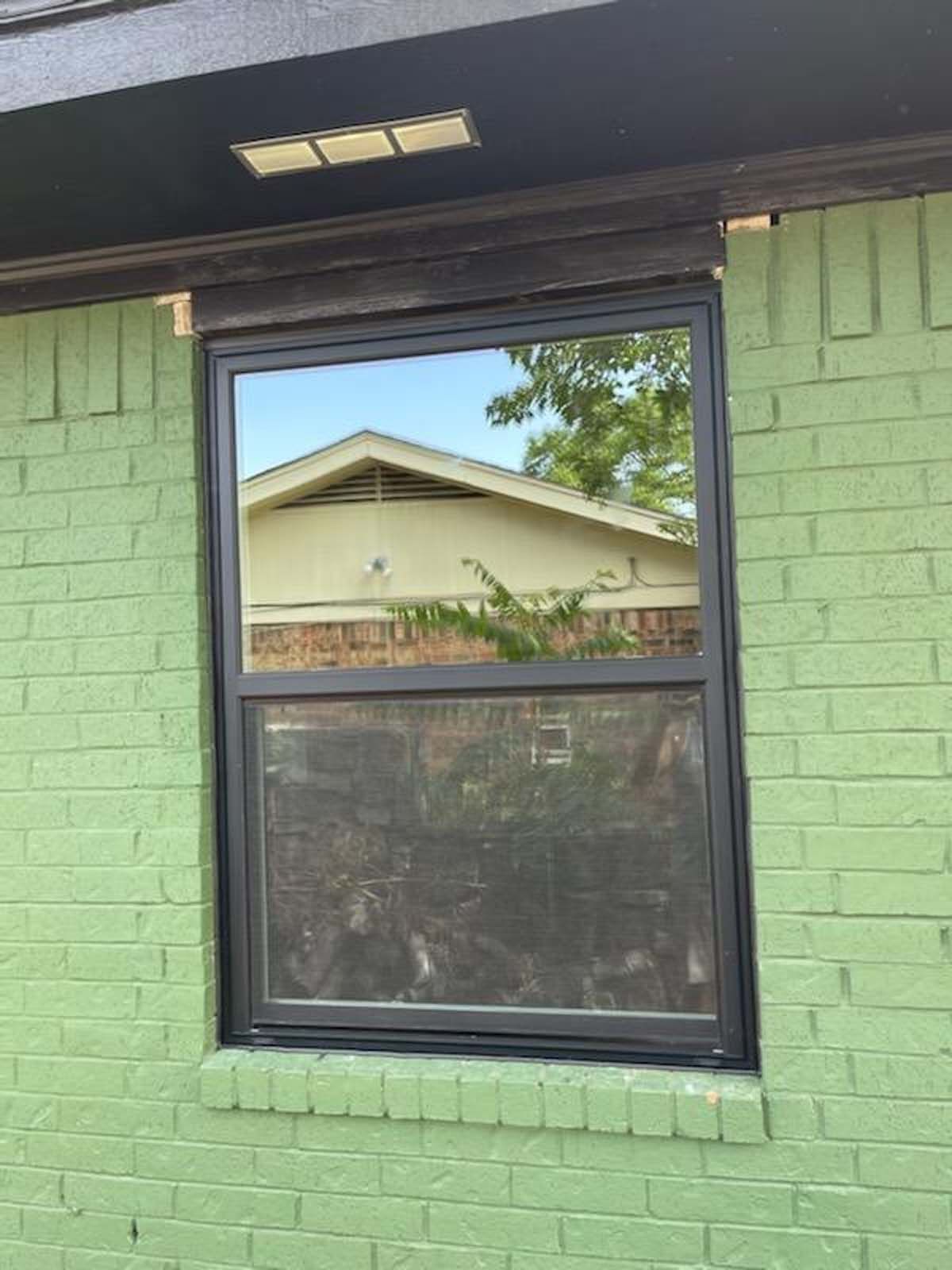 Black-framed window on a green brick wall reflecting a house and trees.
