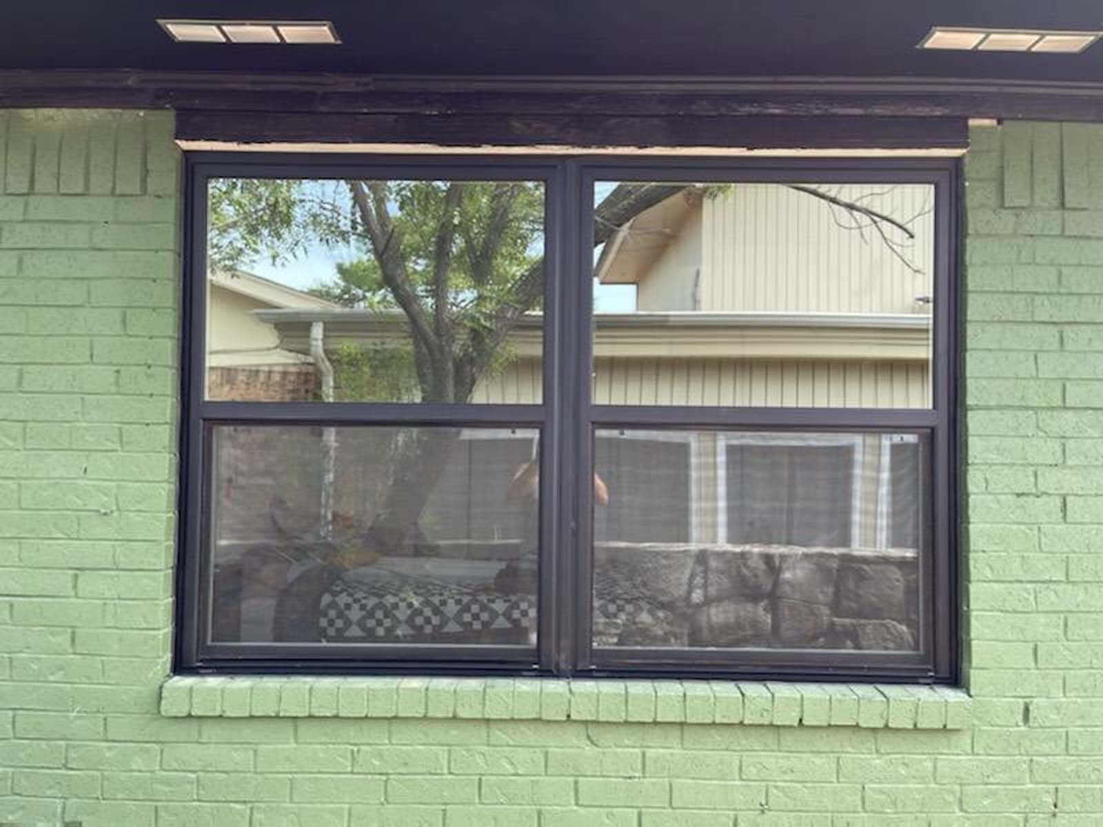 Black-framed window with screen in a green brick wall; reflections of trees and buildings are visible.
