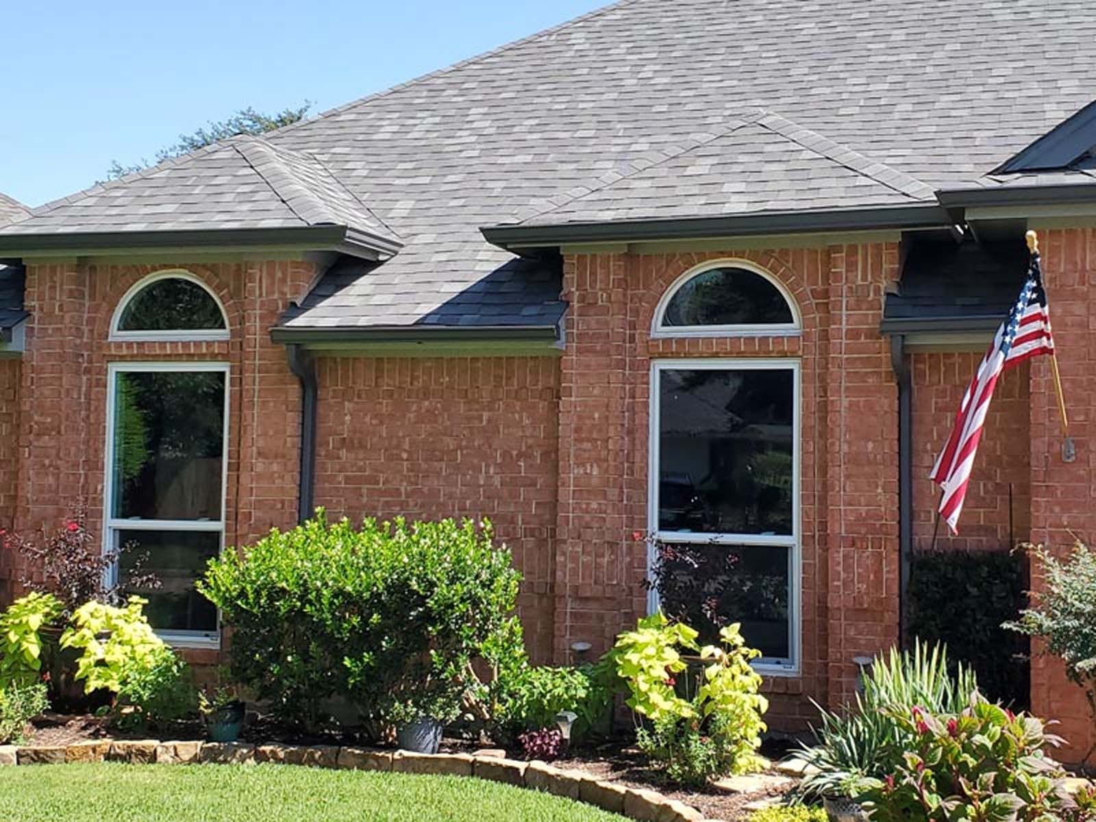 Brick house with two arched-top windows, surrounded by landscaping, under a gray roof. An American flag hangs on the right.