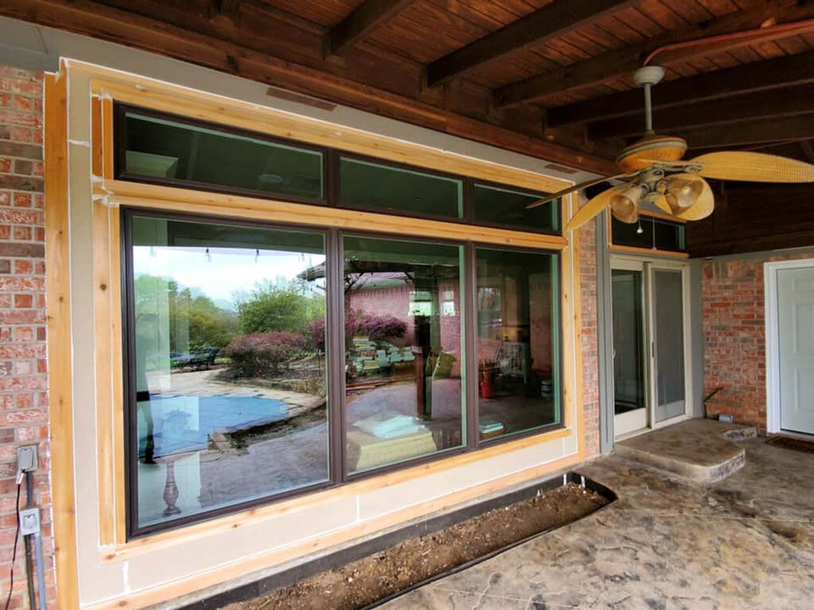 Large windows in a brick home, framed in wood, next to a doorway. A ceiling fan hangs above.