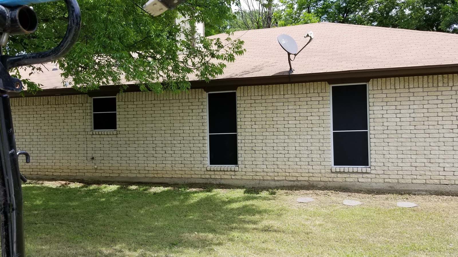 Exterior view of a brick house with dark window screens, satellite dish, and brown roof.
