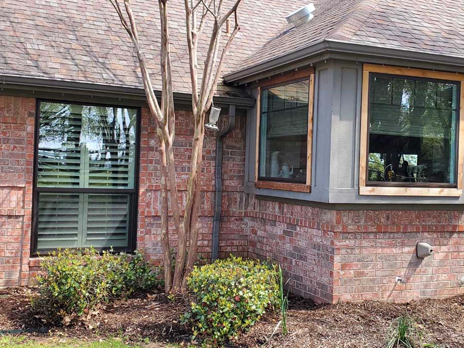 Red brick house with two windows, one with shutters, the other a bay window. A small tree is in front.
