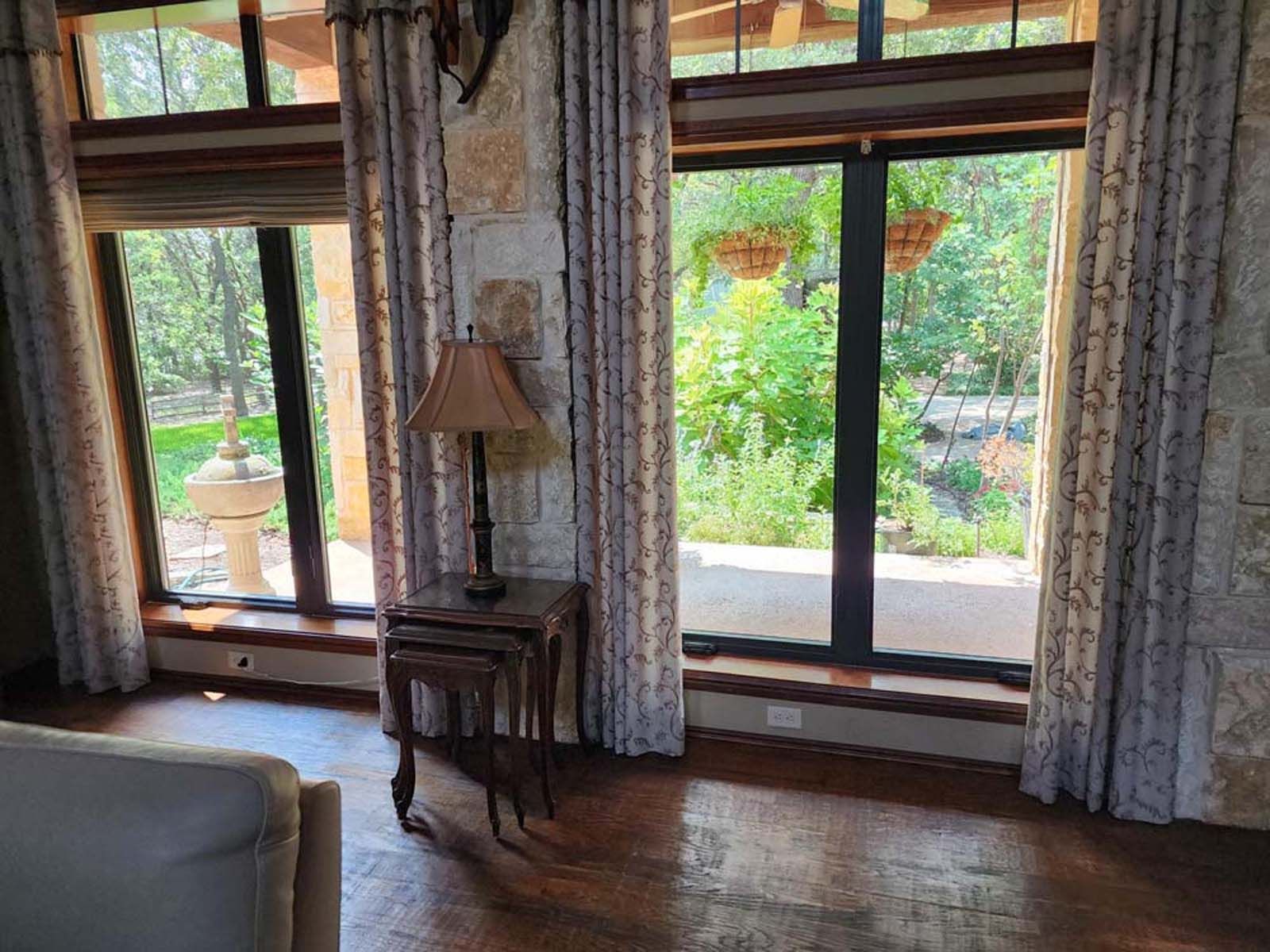Living room with large windows, stone walls, and curtains. A table with a lamp sits between the windows, and greenery is visible outside.