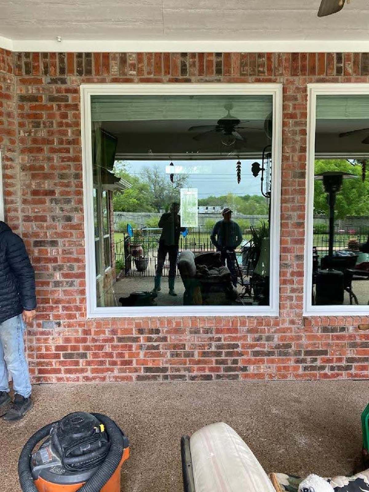 Large window with brick exterior. Two people reflected in the glass.  A vacuum cleaner sits on the patio.