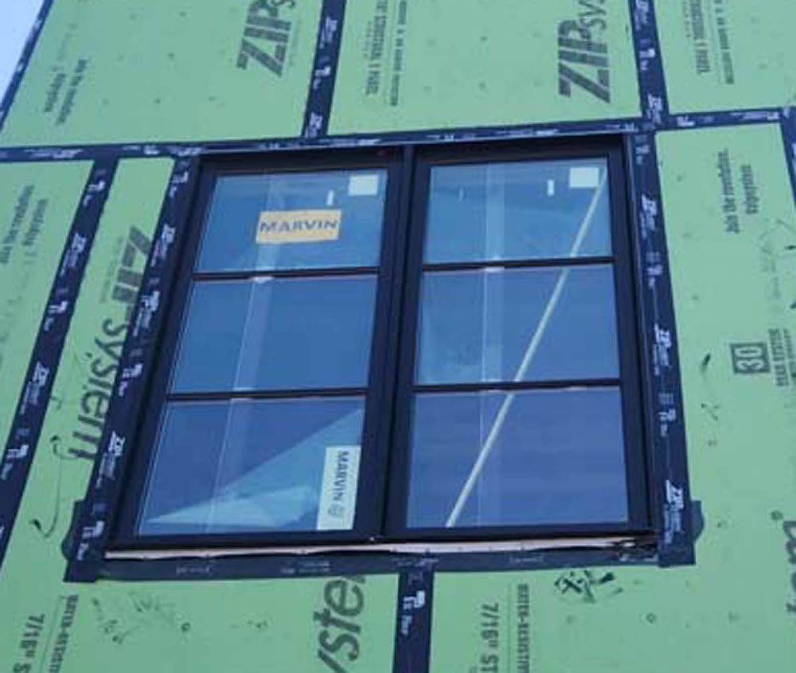 A black-framed window installed on a building exterior, surrounded by green sheathing.