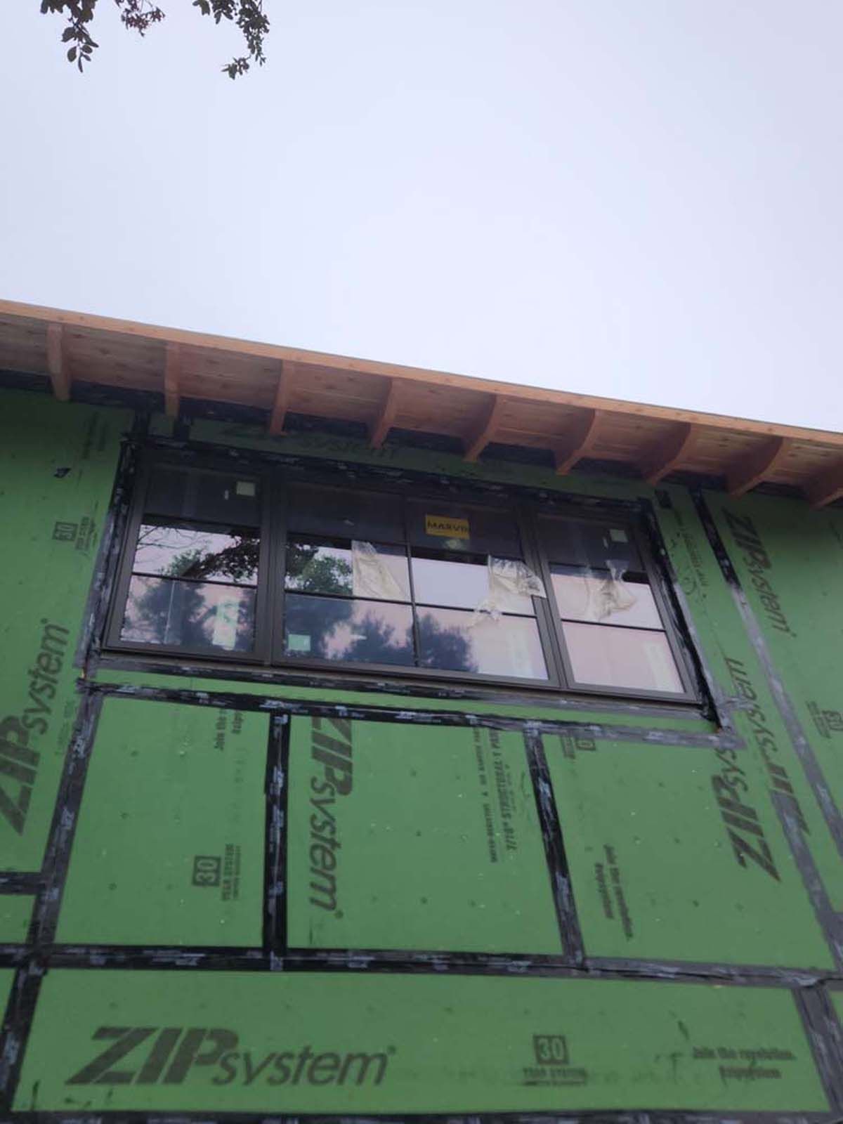 Exterior of a building under construction. A large window is installed in green sheathing. Sky overhead.