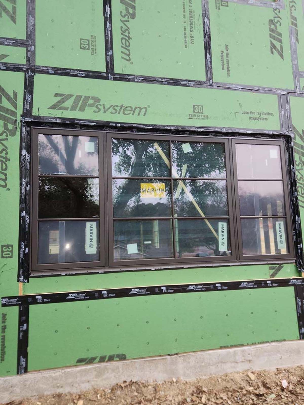 Brown window installed on a new construction wall with green sheathing and black sealant tape.