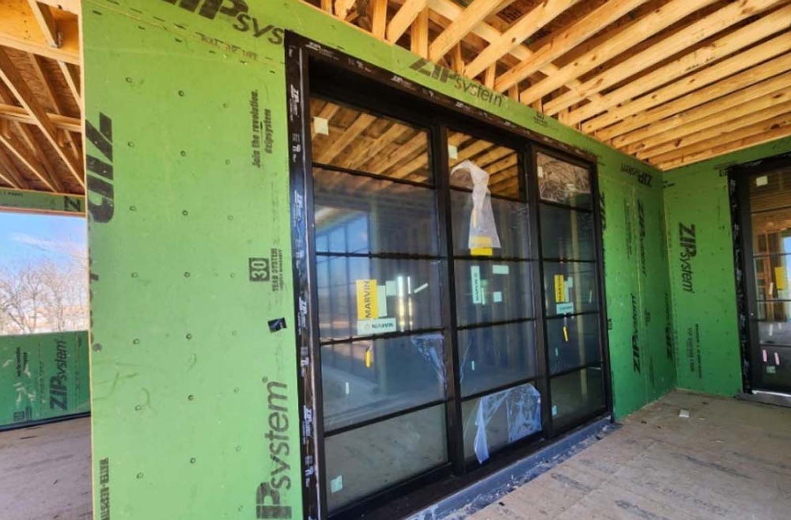 Large black-framed windows installed in a new construction home. Green sheathing and exposed wood framing visible.