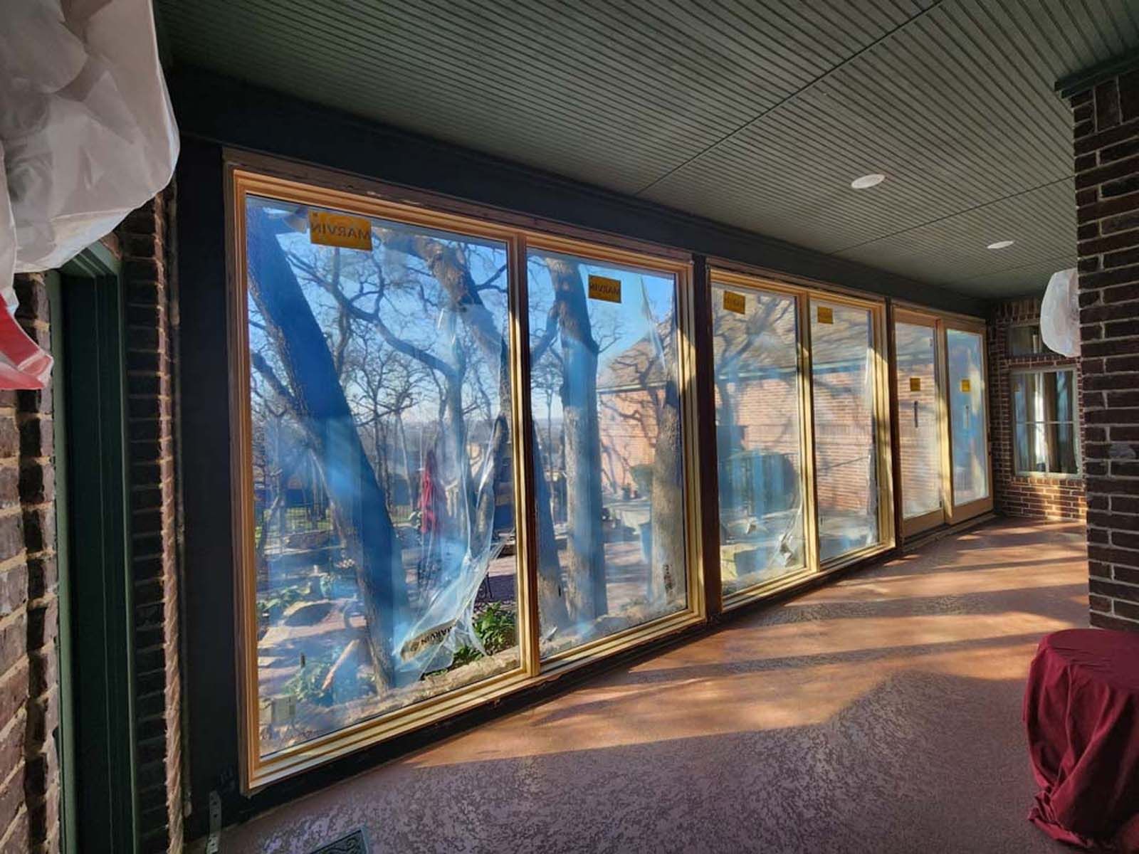 A row of large windows with gold frames and protective covering, overlooking trees and a green outdoor area.