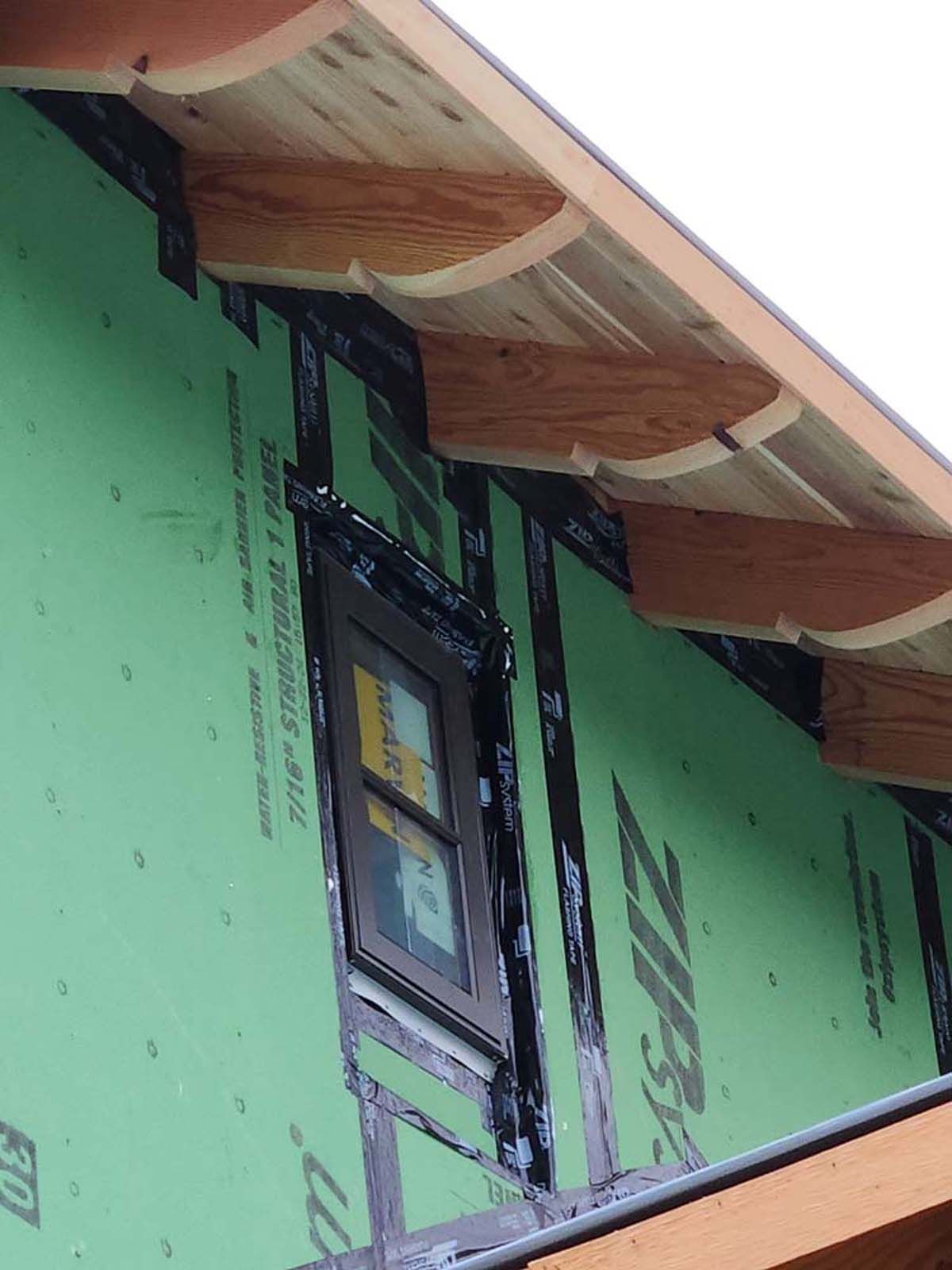 Exterior of a building under construction, showing a window, green sheathing, and wooden rafters.