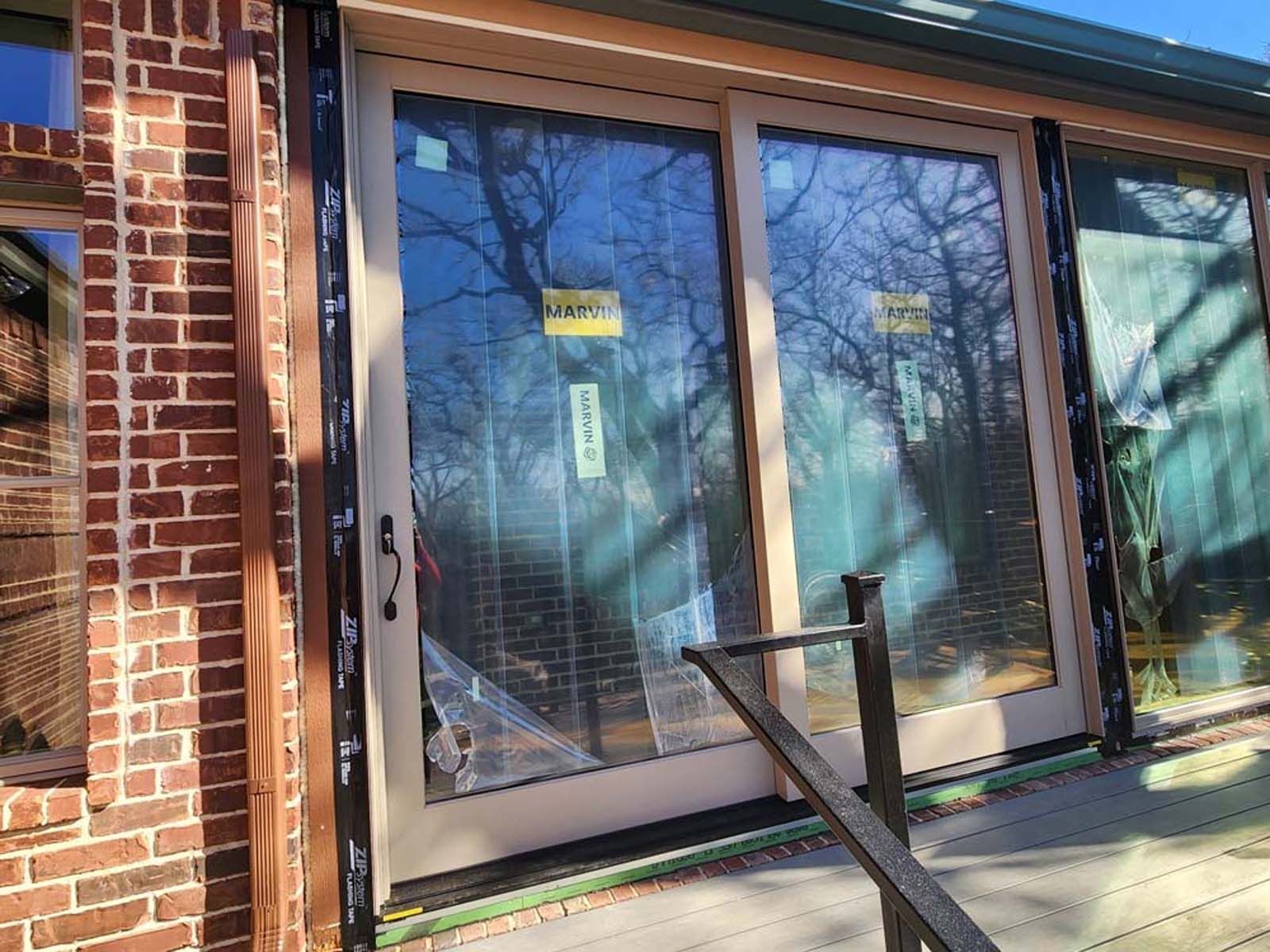 Sliding glass door, beige frame, black trim, on brick building, partially covered with plastic.