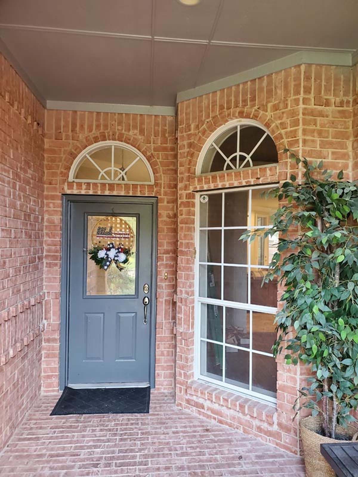 Gray front door with arched window above, next to a large window and brick exterior.