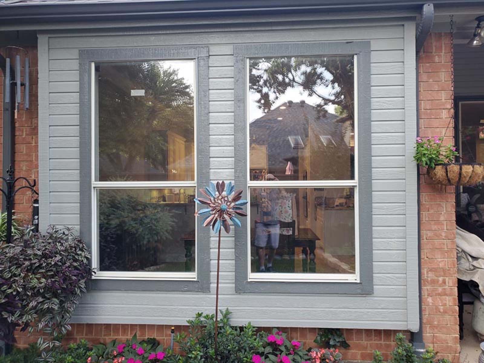 Two large windows with gray trim reflect a yard; a brick wall and plants are visible.