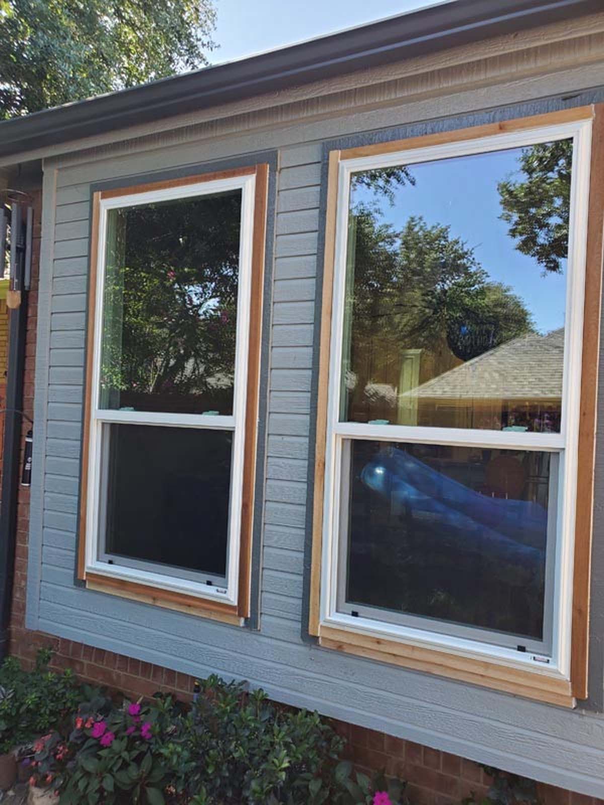 Two double-hung windows with white frames, surrounded by wood trim, on a gray-sided building.