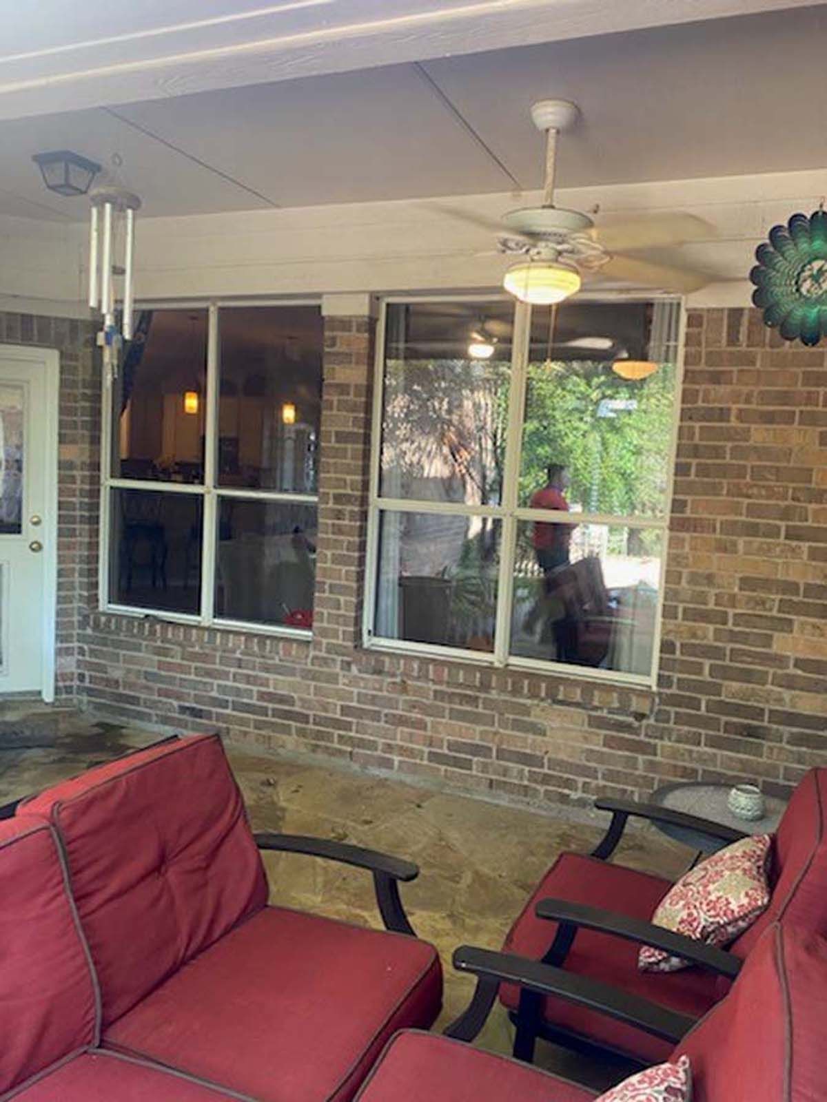 Patio with red cushions, brick wall, two windows, wind chimes, ceiling fan, and outdoor seating.