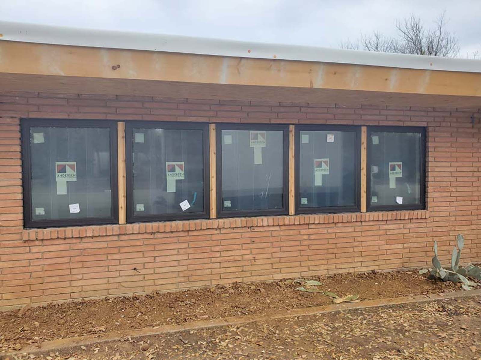 Brick building exterior with four black-framed windows; dirt and small plants in the foreground.