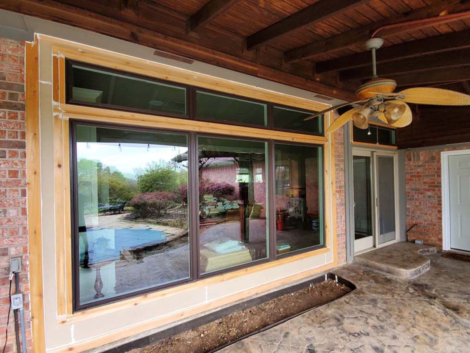 Large windows on a patio overlook a backyard with pool. Brown window frames, brick walls, ceiling fan.