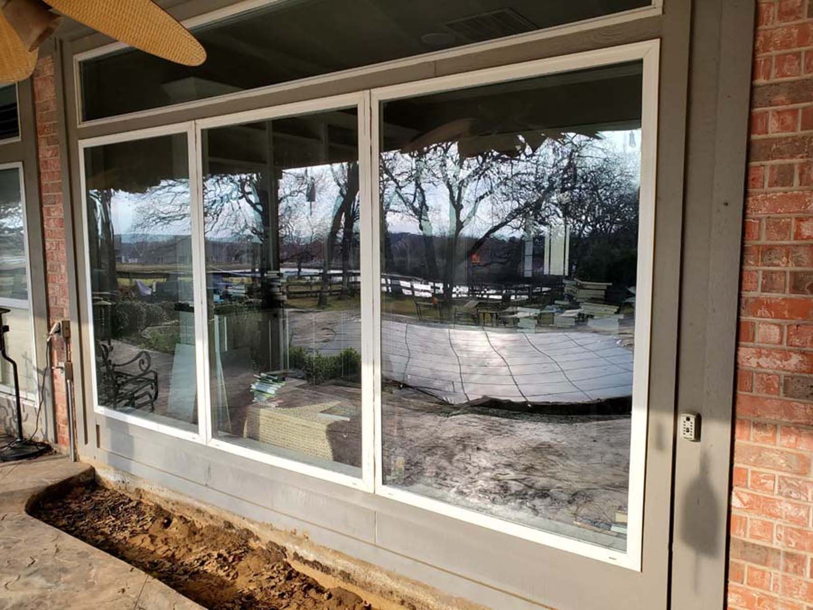 Glass sliding doors with white frames reflect a backyard scene with trees and a patio.
