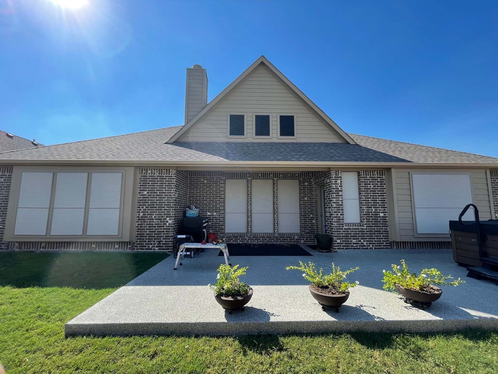 Backyard of a house with a concrete patio, brick exterior, and potted plants under a clear blue sky.