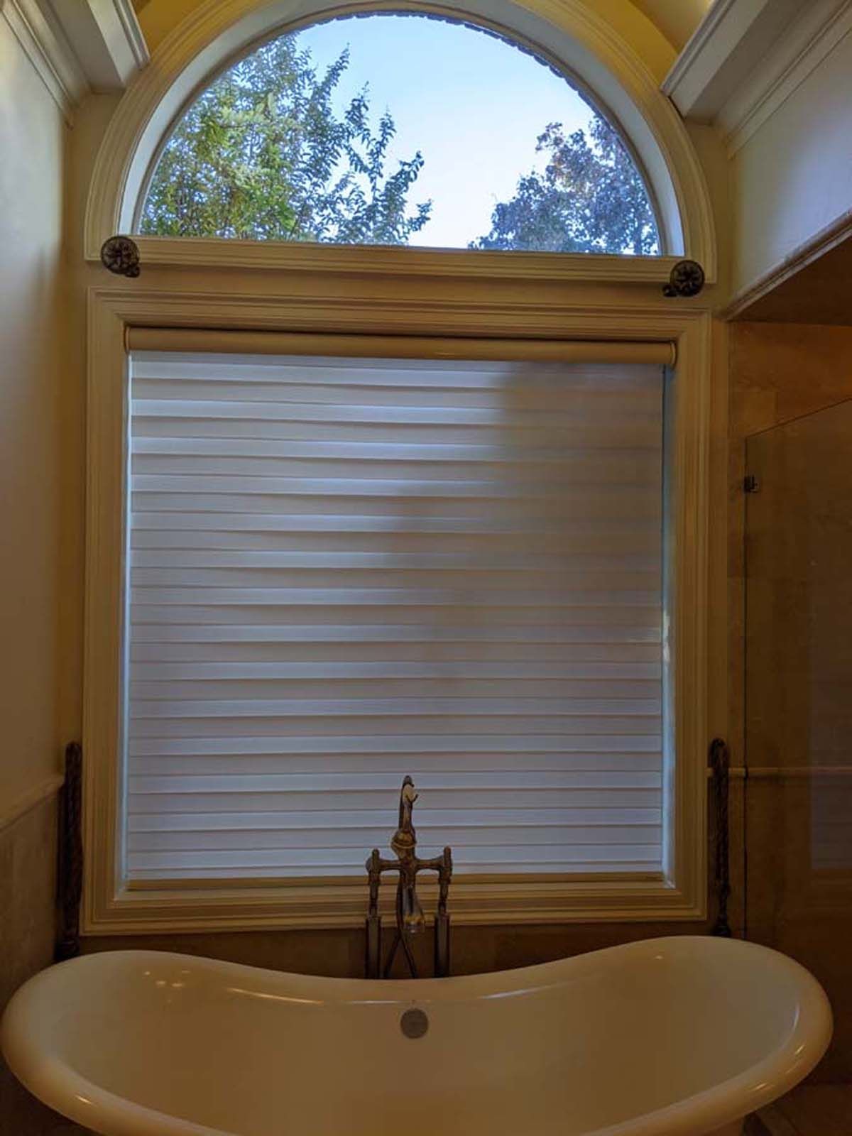 Bathroom with a white bathtub below a window with a closed blind, the top is an arch shape.