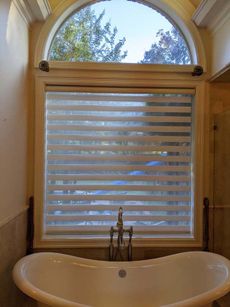 Bathroom window with white blinds above a bathtub. Arched window above displays trees and sky.