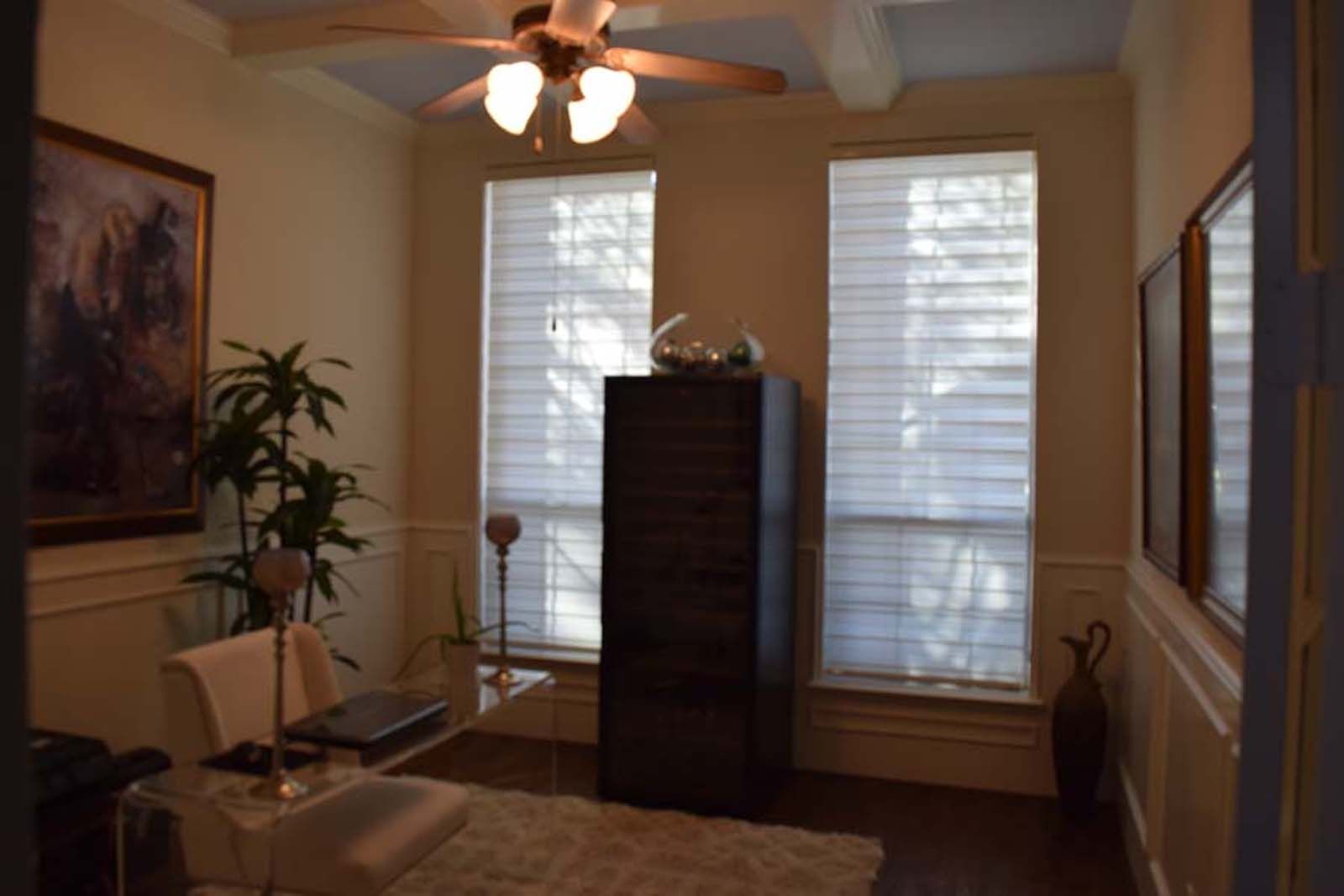 Office with windows, dark storage cabinet, art on walls, ceiling fan, and desk.