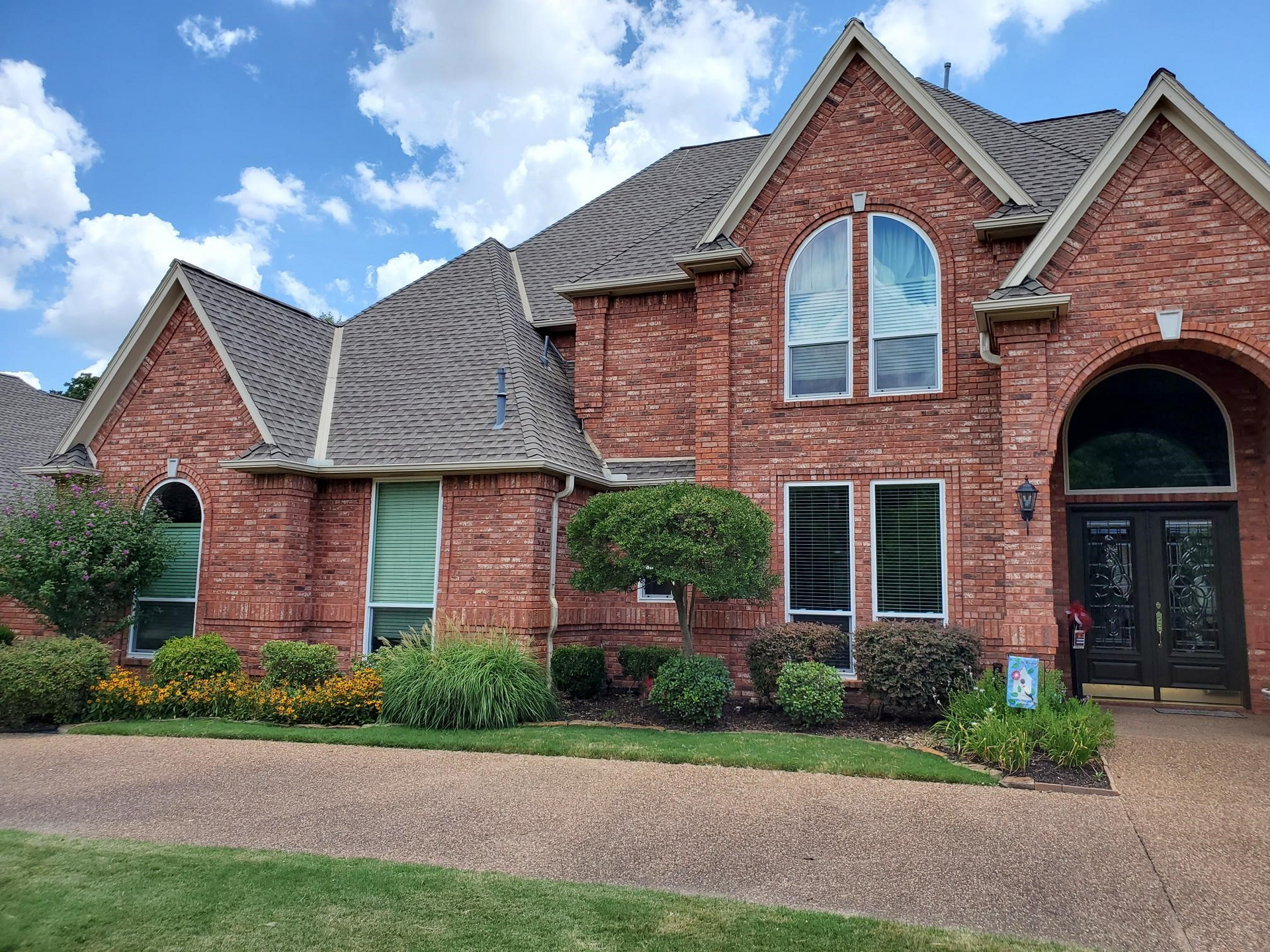 Red brick house with dark roof, arched entry, and landscaping under a blue sky.