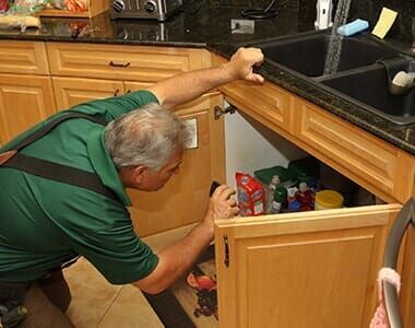 A man in a green shirt is looking under a sink in a kitchen.