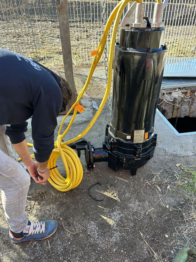 Una persona sta arrotolando un tubo giallo accanto a una grande pompa metallica vicino all'acqua.