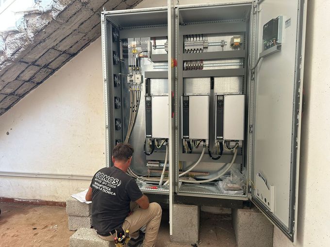 Un tecnico è inginocchiato accanto a un quadro elettrico aperto, intento a lavorare sul cablaggio all'esterno, sotto una scala.