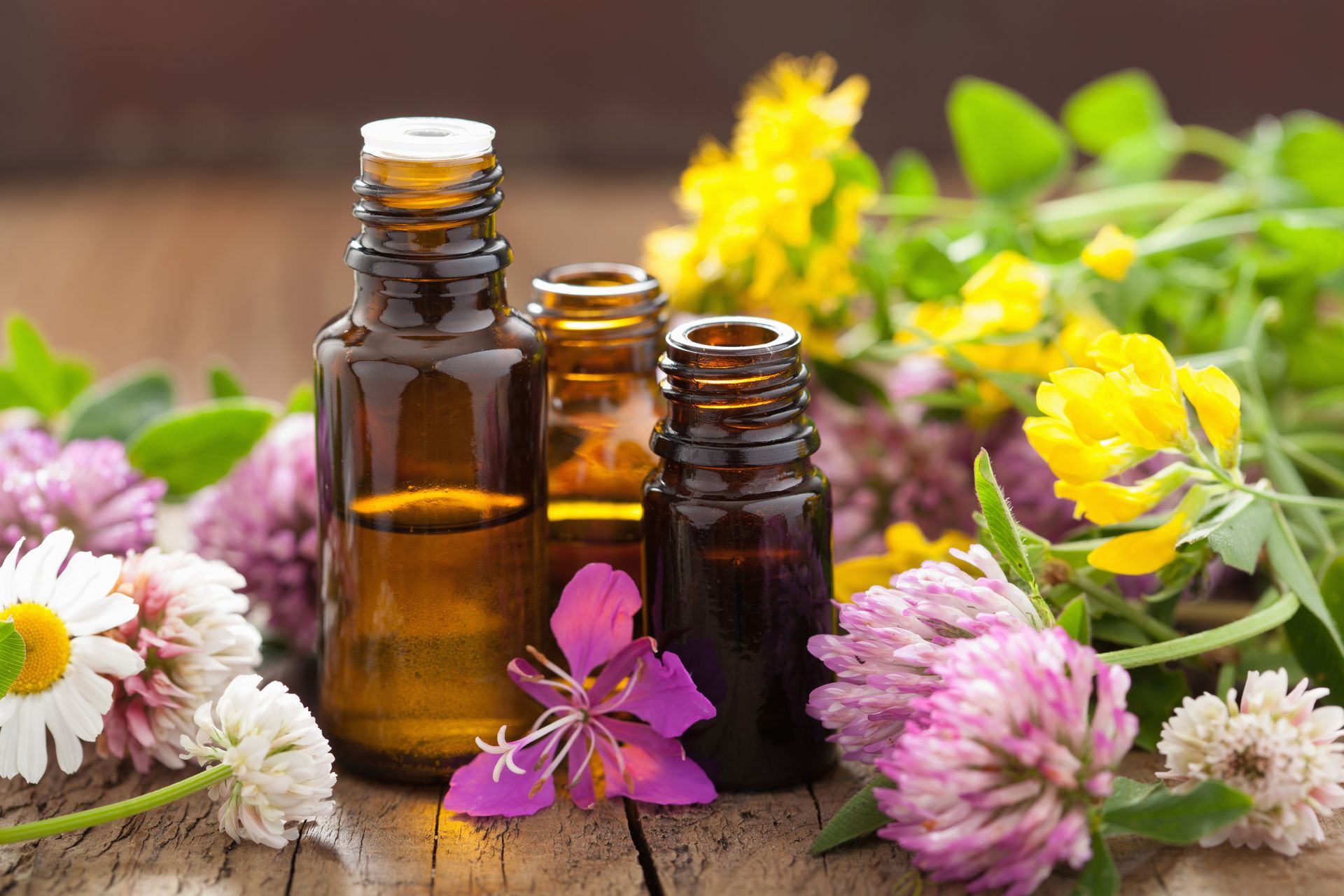 Three bottles of essential oil surrounded by flowers on a wooden table.