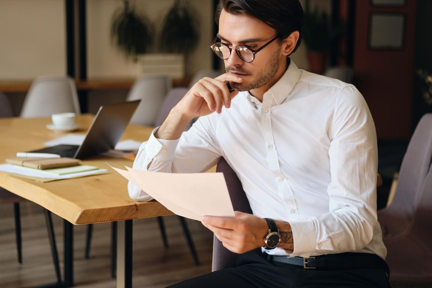 Man reading a document at a table with a laptop in a modern office.