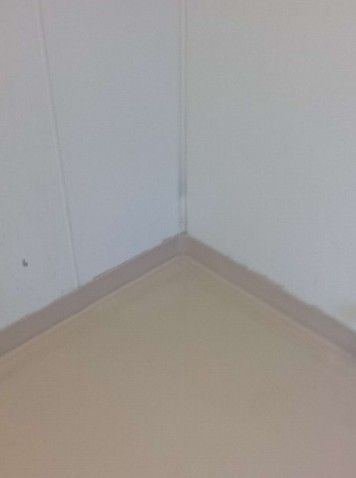 Empty corner of a white room with beige baseboard and light tan floor.