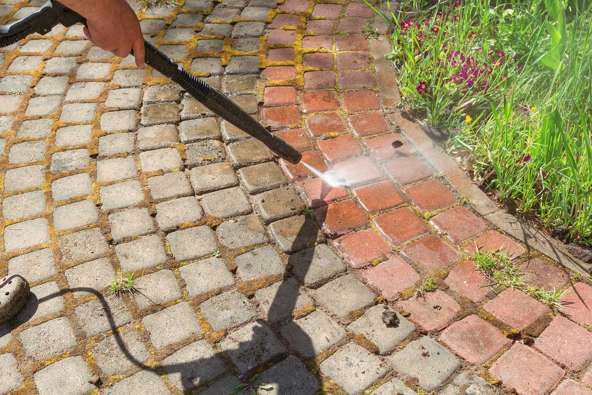 Person pressure-washing a cobblestone walkway beside a garden with flowers and greenery
