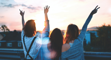 Three women are standing on a balcony with their arms in the air.