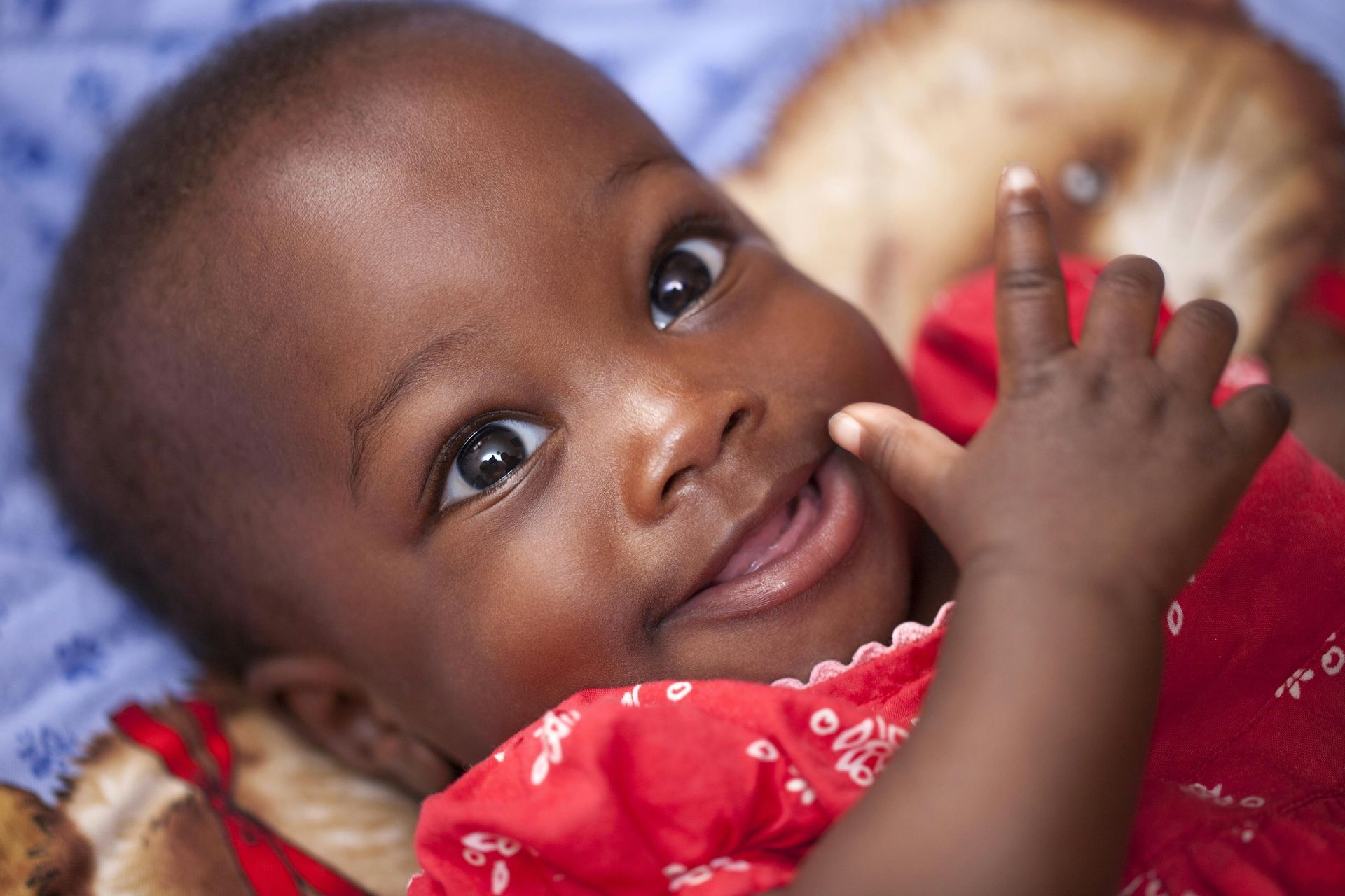 A baby in a red dress is laying on a blue blanket and smiling.