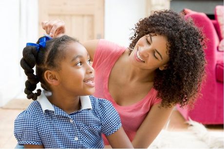 A woman is brushing a little girl 's hair with a blue bow.