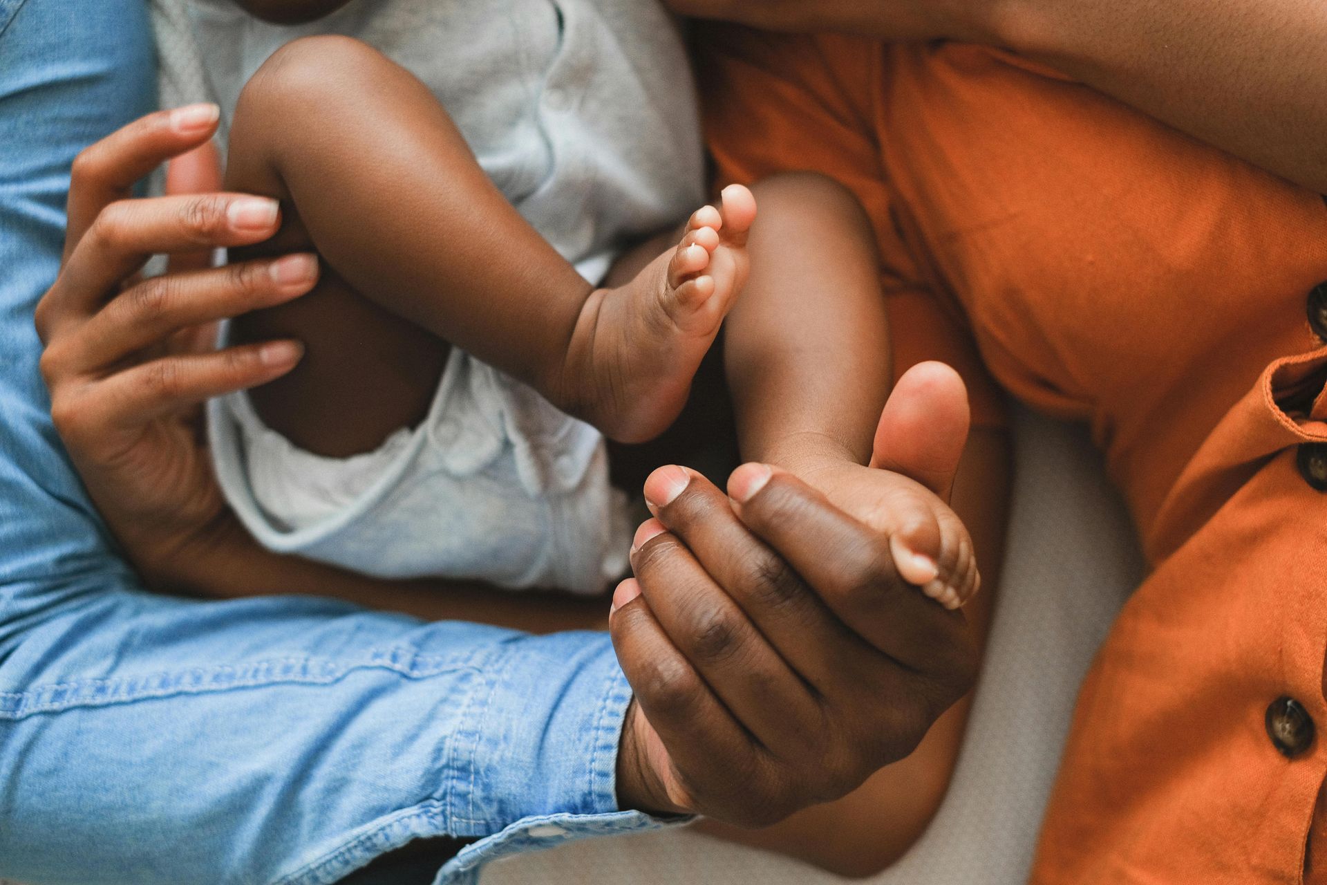 Person holding a baby's foot. The baby is wearing a white onesie. Another person in orange is next to them.