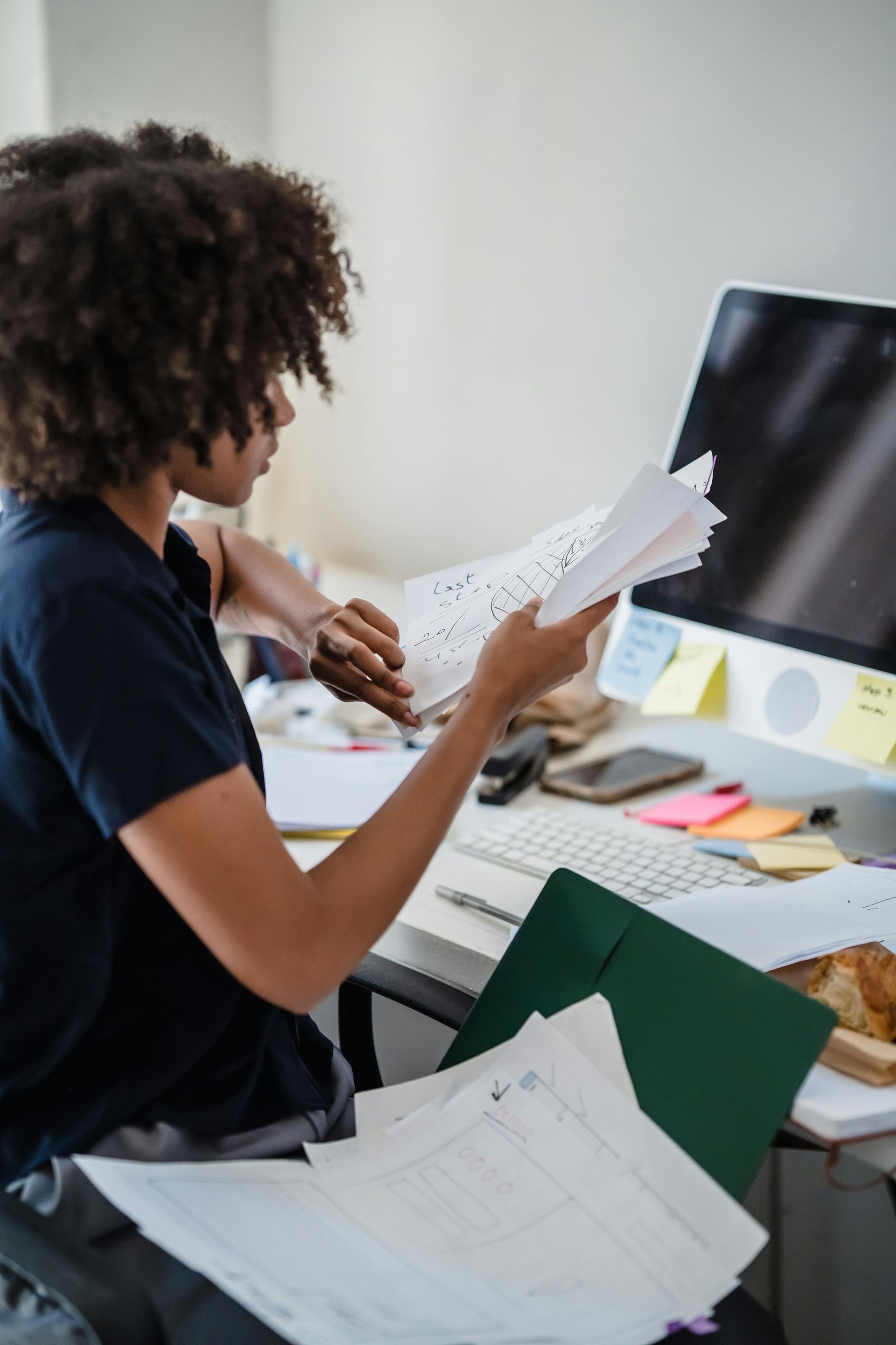 Woman examining papers at a messy desk with a computer and green folder.