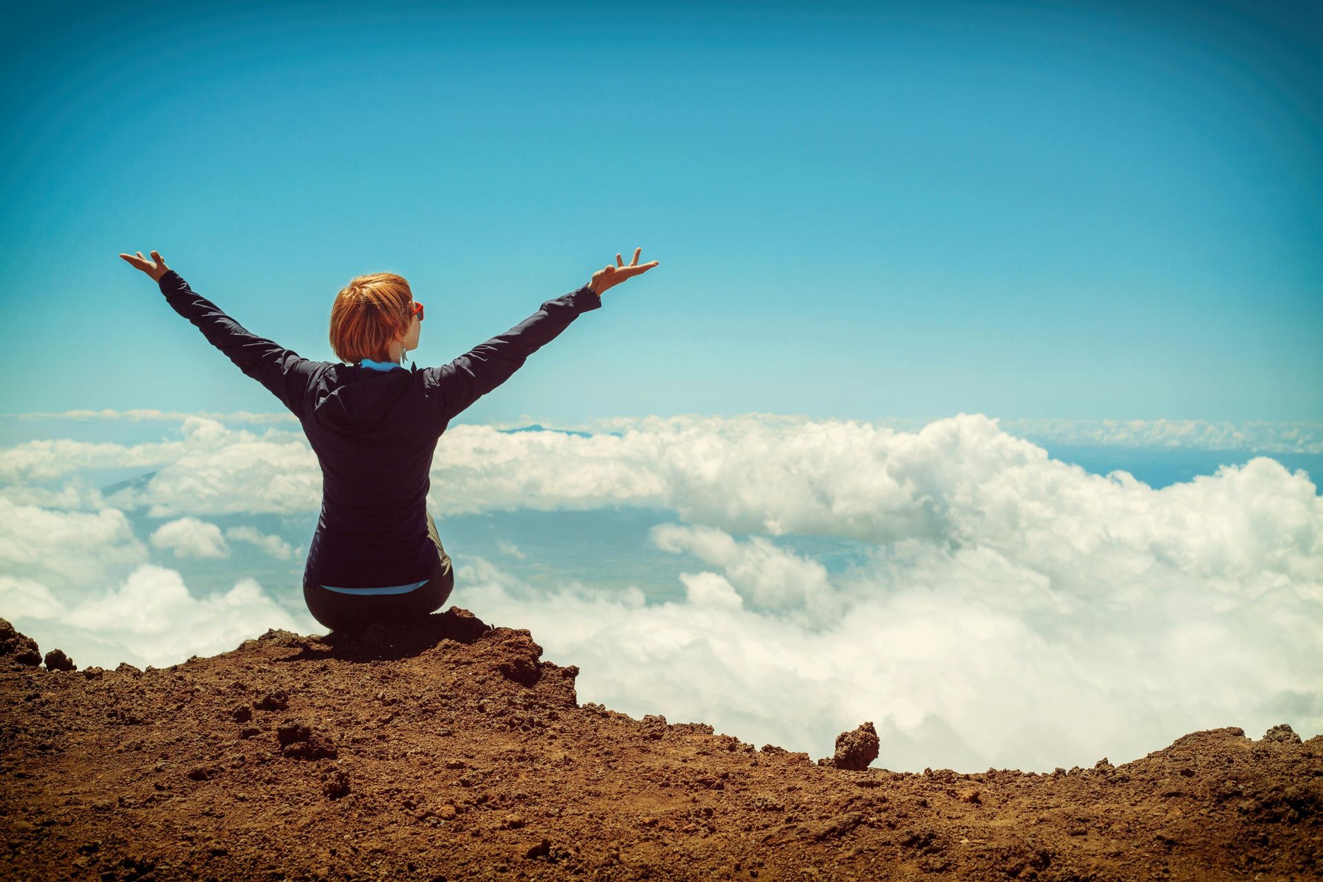 Woman with arms outstretched, seated on mountaintop, gazing at sky above clouds.