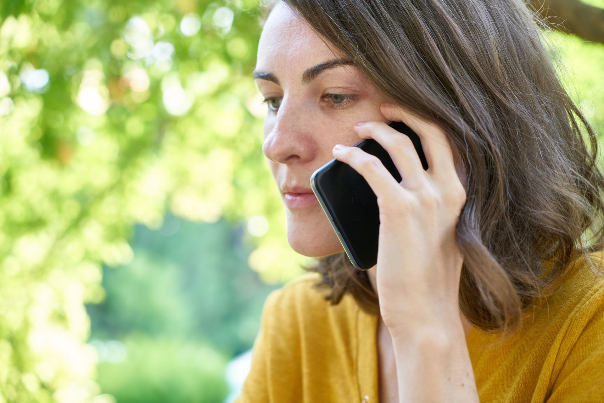 Person in a mustard sweater talking on a smartphone outdoors against a leafy green background