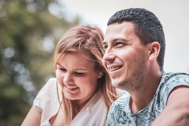 Two people smiling outdoors, leaning close together in soft daylight