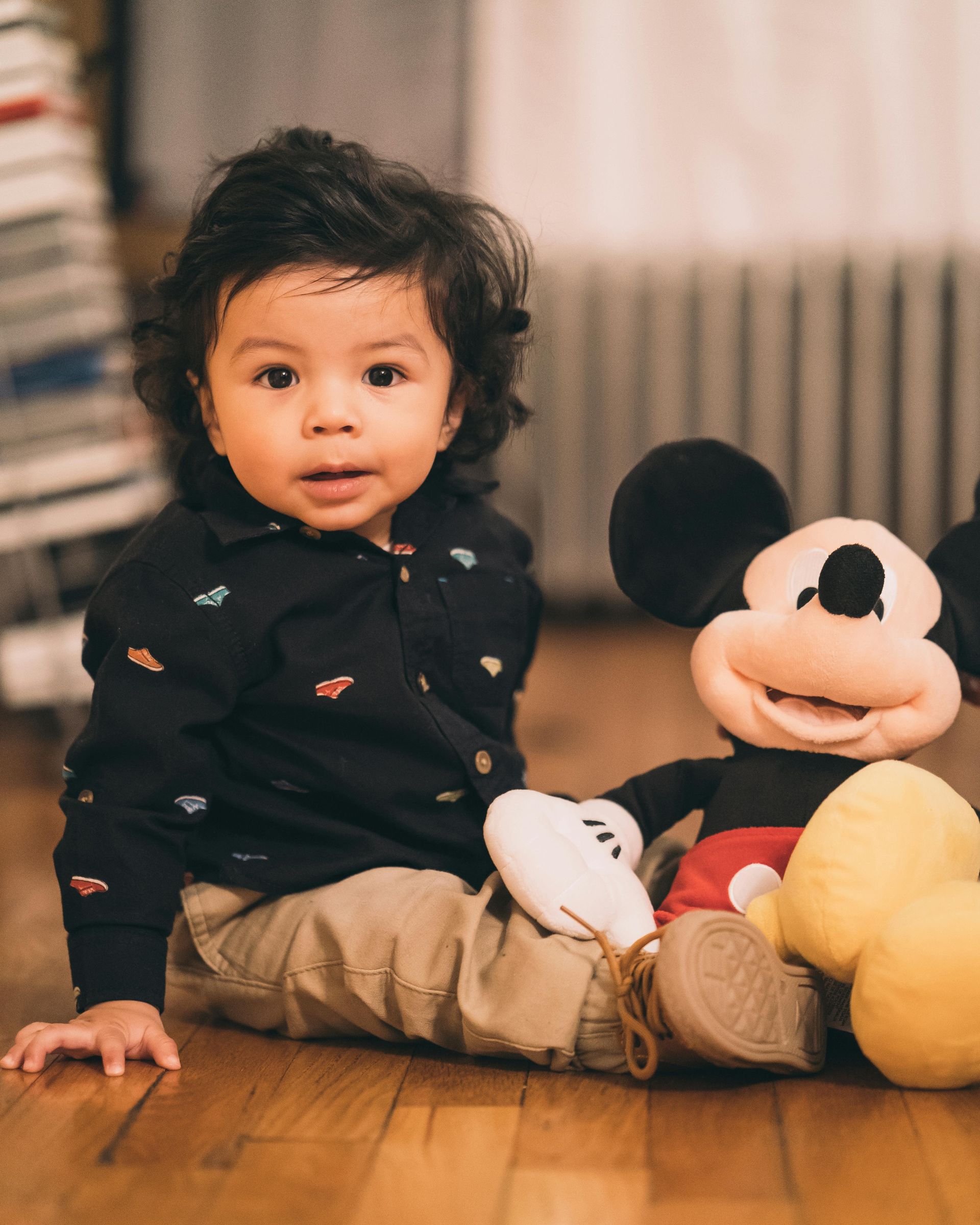 Child sitting on floor next to Mickey Mouse plush. Child has dark hair and is wearing a dark shirt and khaki pants.