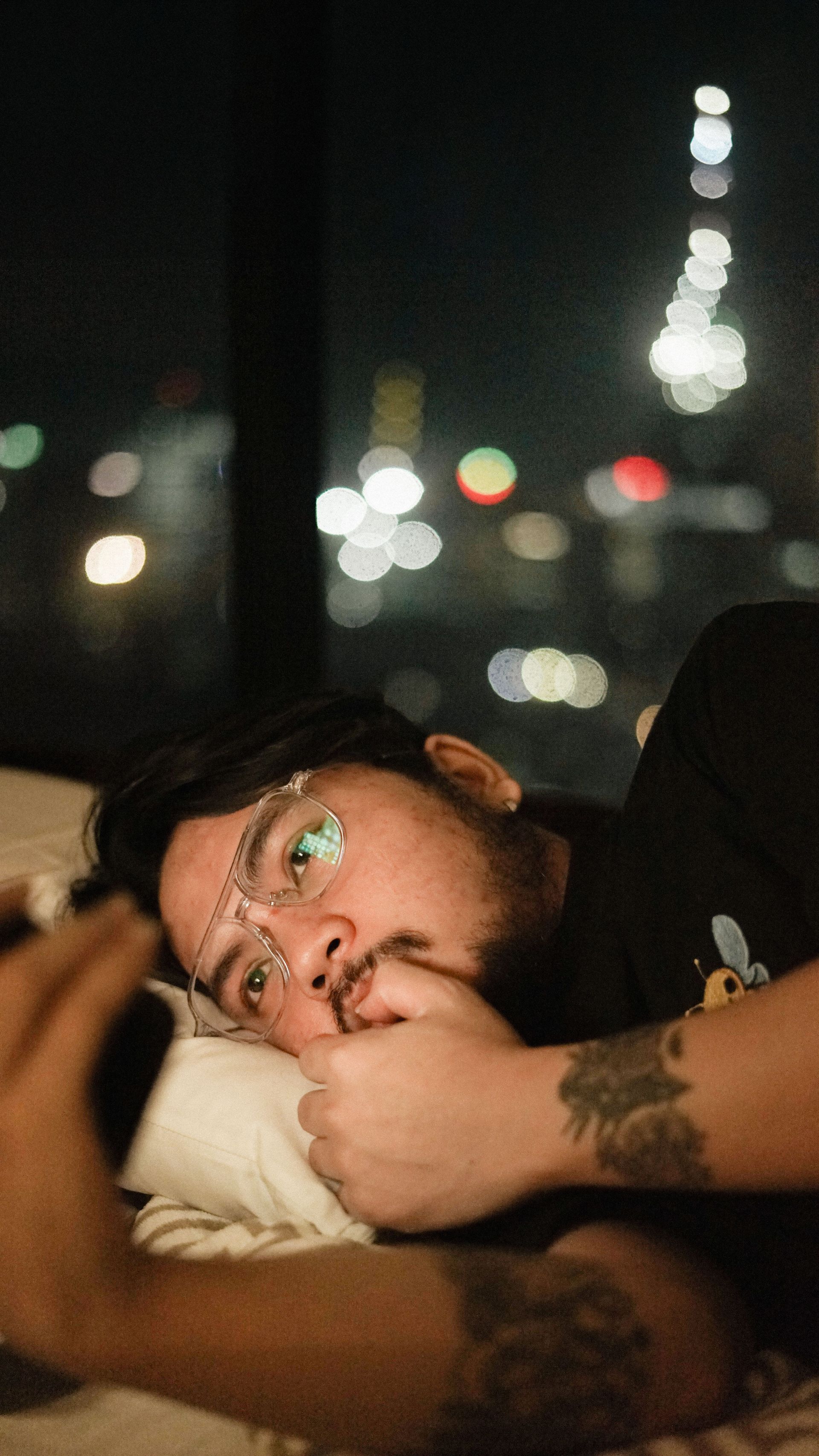 Man lying on a pillow in a dim room, gazing upward with city lights blurred in the background.