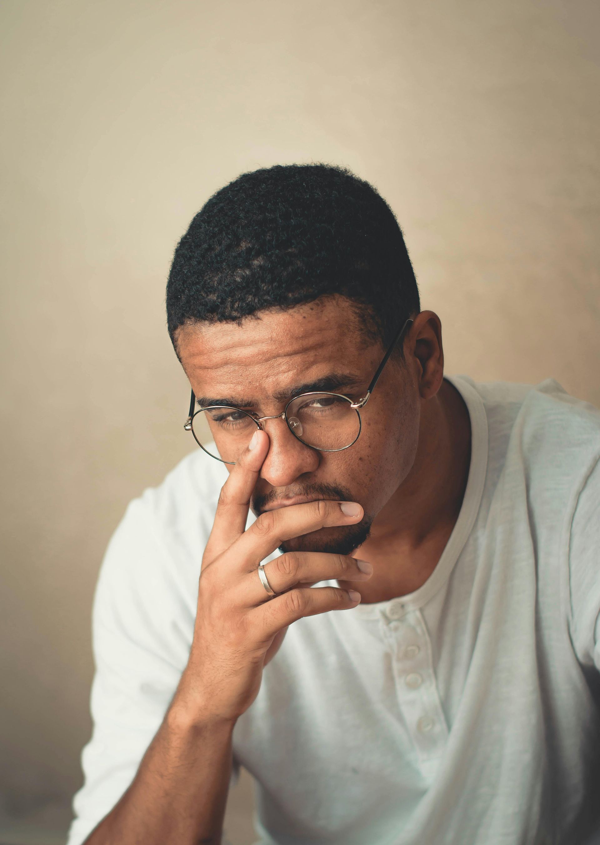 Man with glasses, hand to face, looking thoughtful, wearing a white shirt against a neutral background.