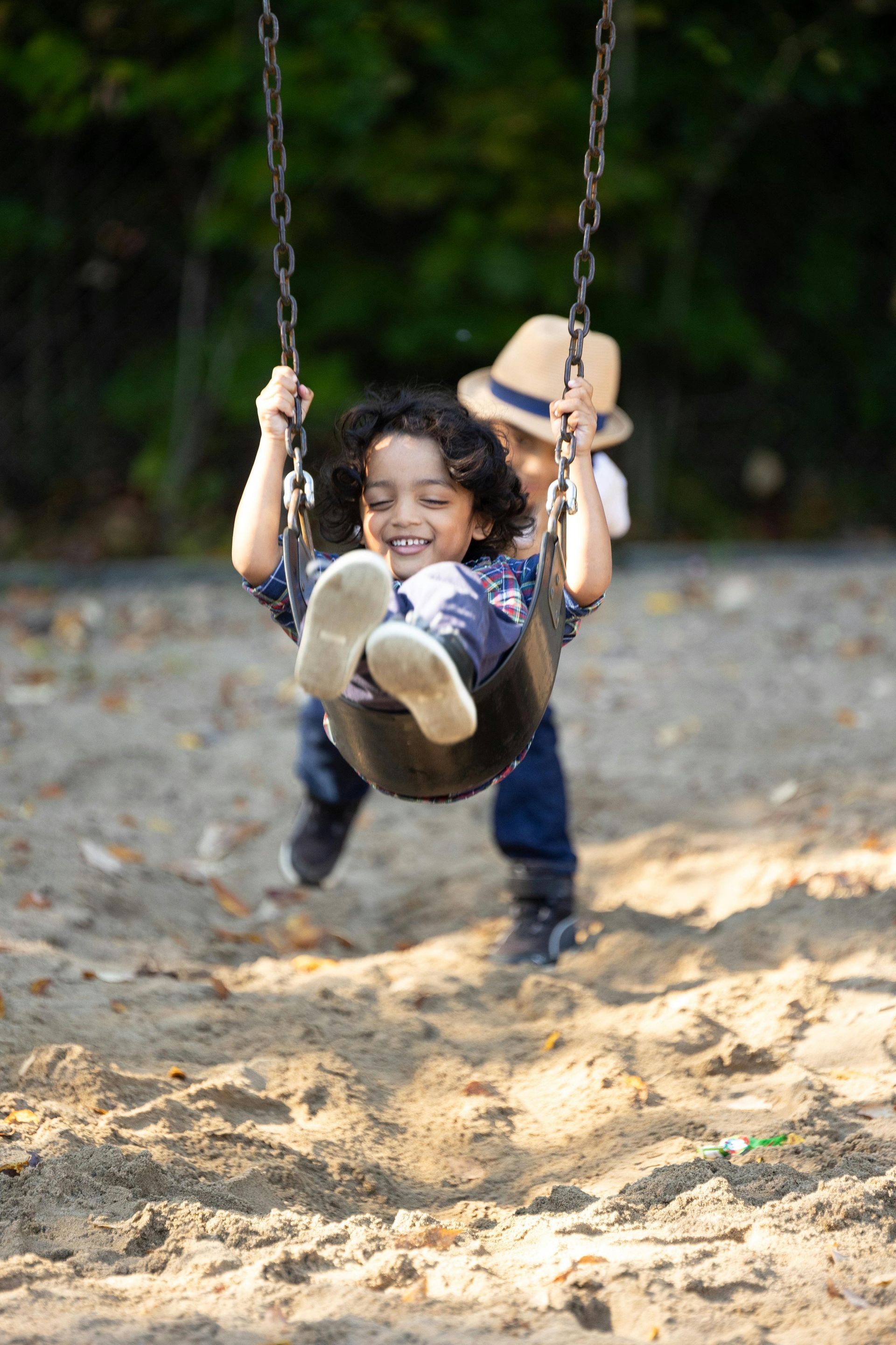 Two children on a swing. One smiles while swinging forward; the other pushes from behind. Set in a sandbox.