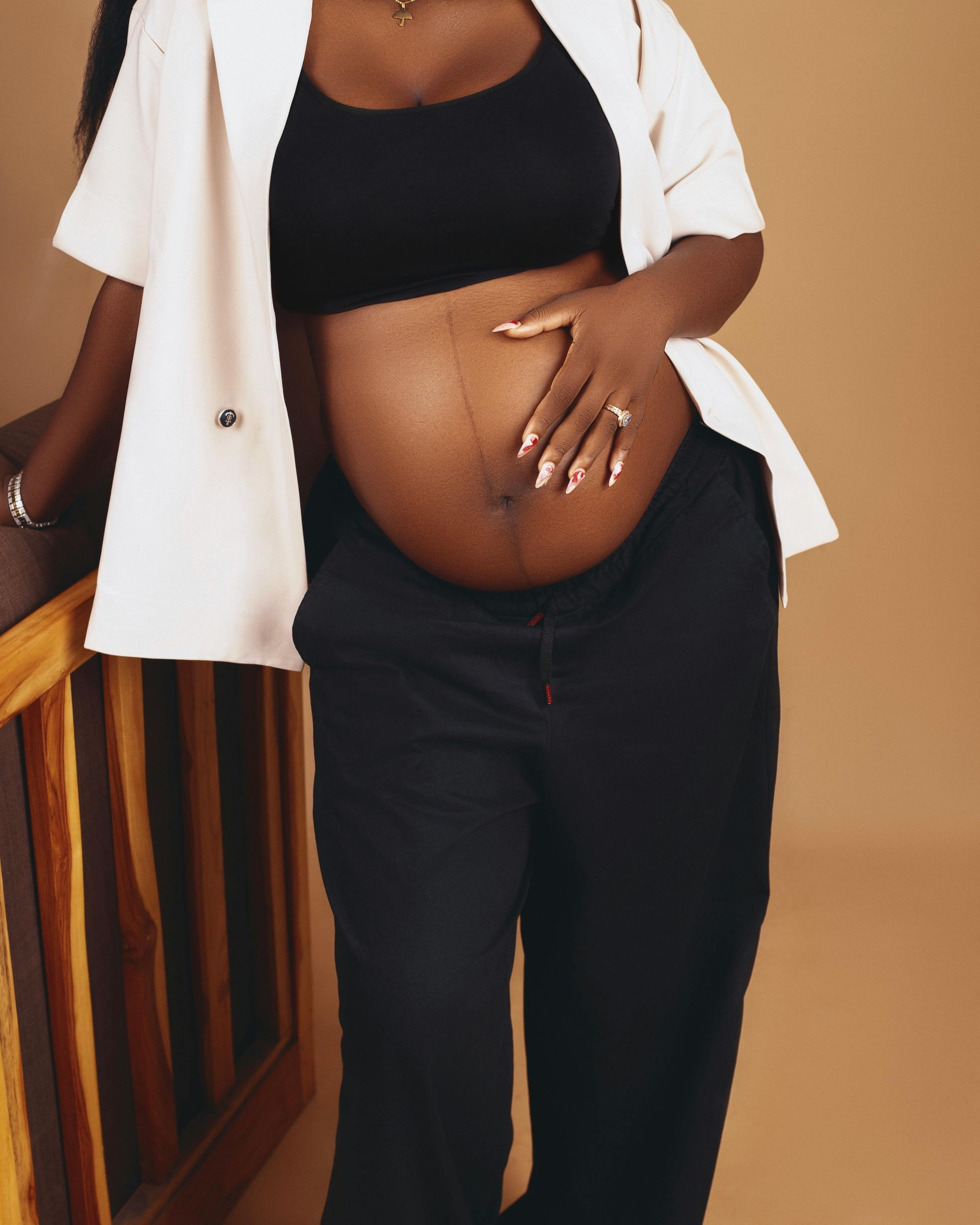 A woman wearing a white blazer and black pants holds their bare, pregnant belly in a studio setting.