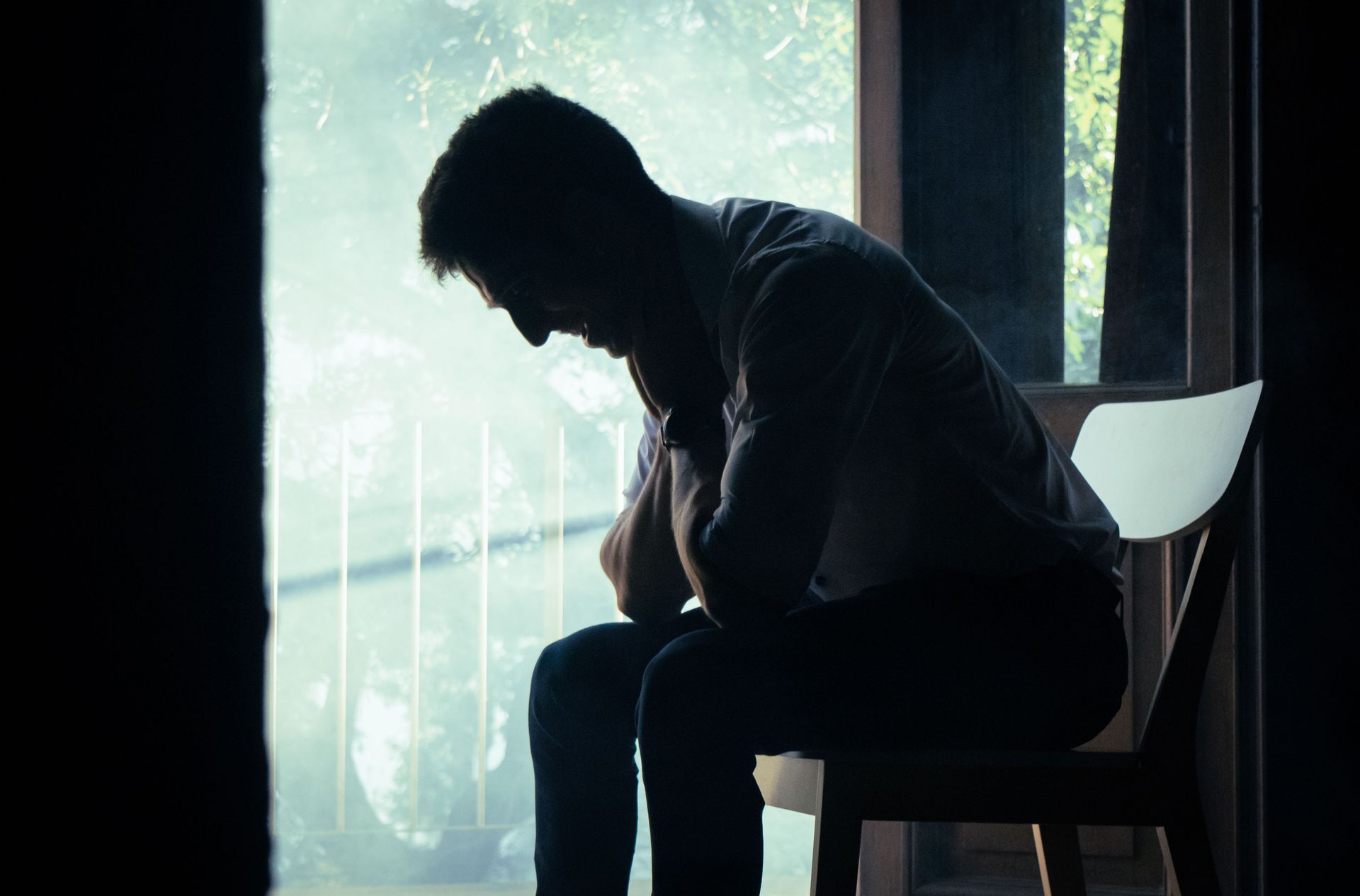 Silhouetted person sitting hunched by a bright window, head bowed in a dark room