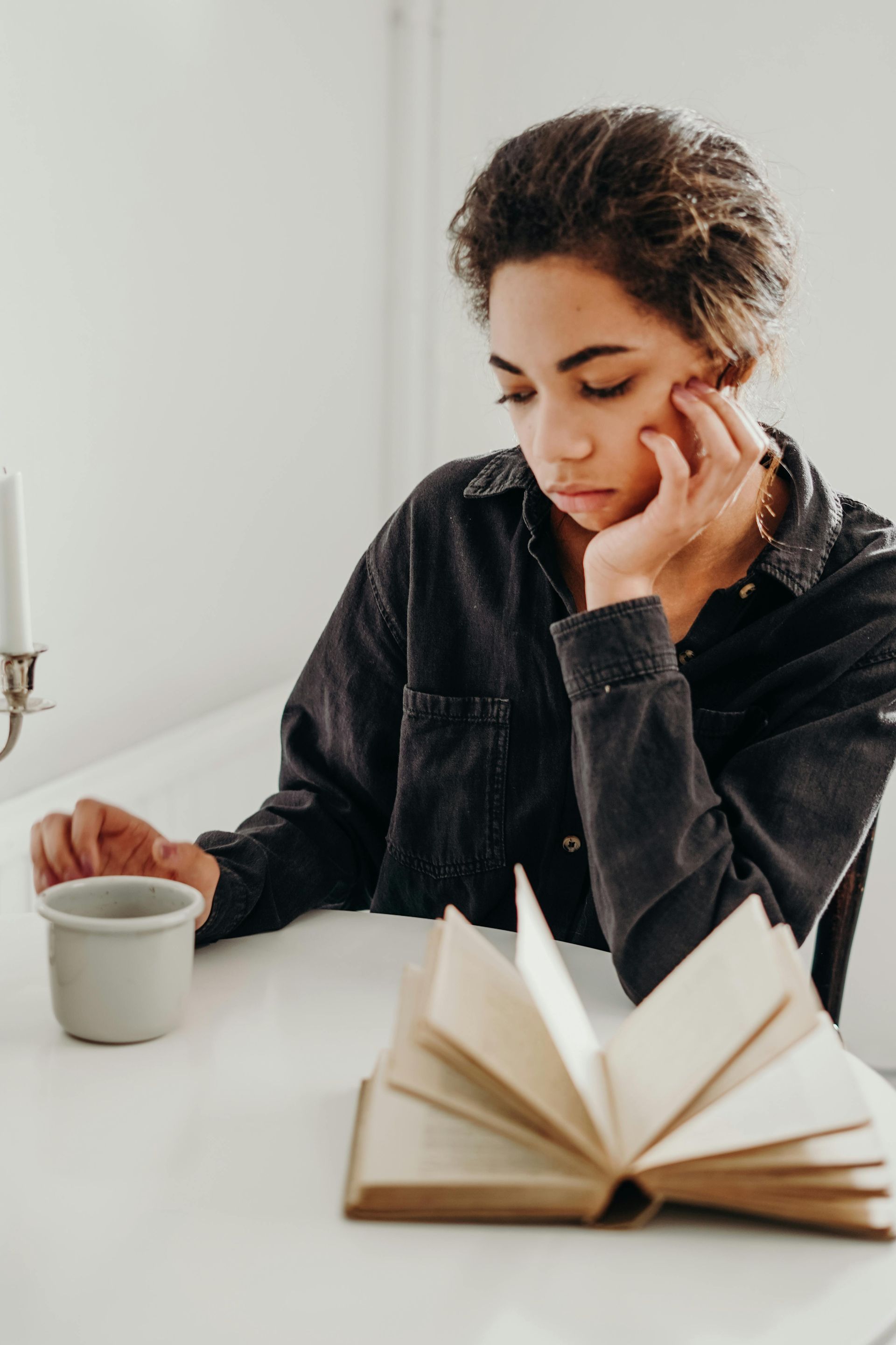 Woman at a table, reading book, resting her chin on her hand, a mug on the table.